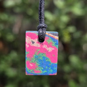 May include: A rectangular pendant with a vibrant, abstract design in pink, blue, and green hues. The pendant is suspended from a black cord, creating a striking contrast. The overall design is eye-catching and unique.