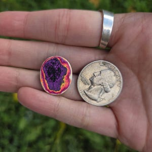 May include: An oval-shaped, multi-colored stone with a dark purple center, surrounded by orange and yellow hues. A silver quarter coin is next to the stone. The stone is approximately 0.75 inches in length.