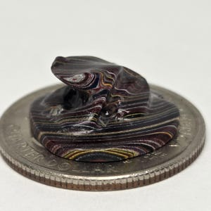 May include: A small, abstract sculpture with a layered, swirled pattern of dark red, black, yellow, and white. The sculpture sits on a quarter coin, showcasing its size. The piece has a unique, organic shape.