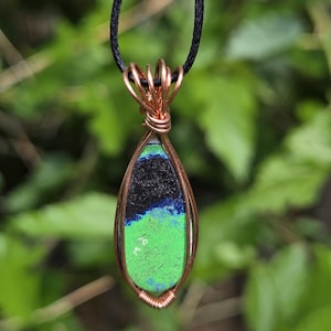 May include: A teardrop-shaped pendant with a black cord. The pendant features a stone with a green, blue, and black pattern, encased in copper wire. The wire is intricately wrapped around the stone, creating a decorative frame.