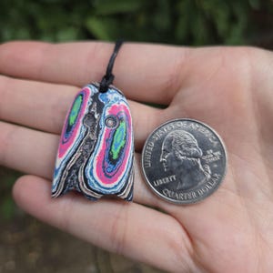 May include: A colorful pendant with a unique pattern of pink, blue, green, and black swirls. The pendant is attached to a black cord. A US quarter coin is next to the pendant for size comparison.