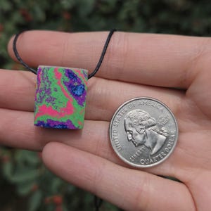 May include: A rectangular pendant with a vibrant, multicolored design of green, pink, blue, and purple. The pendant is suspended from a black cord. A U.S. quarter coin is next to the pendant for size comparison.