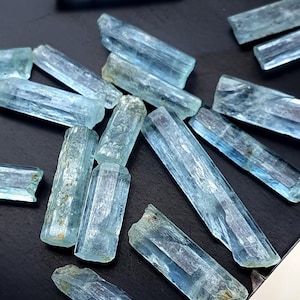 Może przedstawiać: Kolekcja jasnoniebieskich kryształów akwamarynu w czarnym pojemniku. Kryształy są długie i smukłe, o gładkiej, wypolerowanej powierzchni.