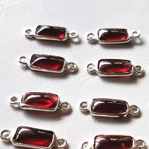 May include: A set of 16 silver-toned connector charms with red gemstone accents. Each charm features a rectangular gemstone with a smooth, polished finish. The charms have two loops on either side, allowing them to be easily attached to jewelry pieces.