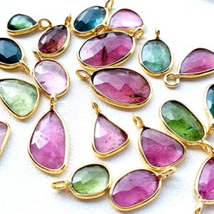 Puede incluir: Una colección de colgantes de piedras preciosas en varias formas y colores, incluyendo rosa, verde y azul. Cada colgante tiene un bisel dorado y un pequeño lazo para colgar. Las piedras preciosas están facetadas y tienen un aspecto pulido.