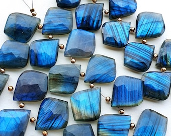 Op de afbeelding: Een verzameling gefacetteerde labradoriet edelsteen kralen in blauwe en grijze tinten, geregen met goudkleurige metalen kralen. De edelstenen hebben een gepolijste, iriserende afwerking en zijn gerangschikt op een wit oppervlak.