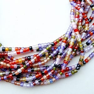 Puede incluir: Primer plano de un collar de múltiples hilos compuesto por pequeñas cuentas de colores. Las cuentas son de varios colores, incluyendo rojo, morado, negro y amarillo. El collar se muestra sobre un fondo blanco.