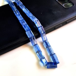 May include: A strand of light blue gemstone beads, each bead is rectangular and faceted. The beads are strung on a clear thread and are lying on a black surface.