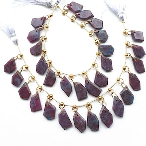 Pode incluir: Fios de contas de pedras preciosas em tons de bordô e cinza azulado. As contas têm formato irregular com corte facetado, enfiadas em fio dourado com pequenas contas douradas. Tassels brancos anexados no topo.