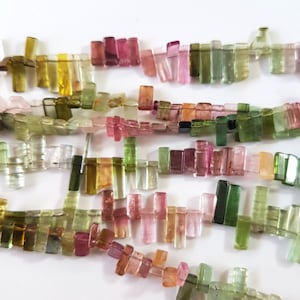 Könnte beinhalten: Eine Strähne aus mehrfarbigen Turmalinperlen. Die Perlen sind facettiert und haben eine rechteckige Form. Die Farben der Perlen umfassen Pink, Grün und Gelb.