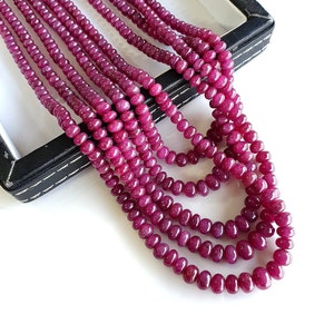 Può includere: Un filo di perle di rubino rosa intenso, con una forma rotonda e liscia. Le perle sono infilate su un filo trasparente e sono esposte su uno sfondo nero.