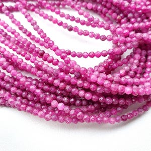 Könnte beinhalten: Nahaufnahme von mehreren Strängen kleiner, runder, leuchtend rosa Edelsteinperlen. Die Perlen sind facettiert und aufgefädelt. Die Perlen haben ein poliertes Aussehen. Der Hintergrund ist weiß.