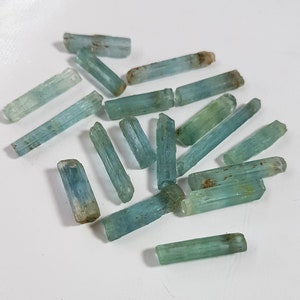 May include: A collection of raw aquamarine crystals, each with a distinct, elongated shape and a light blue-green color. The crystals are arranged on a white surface.