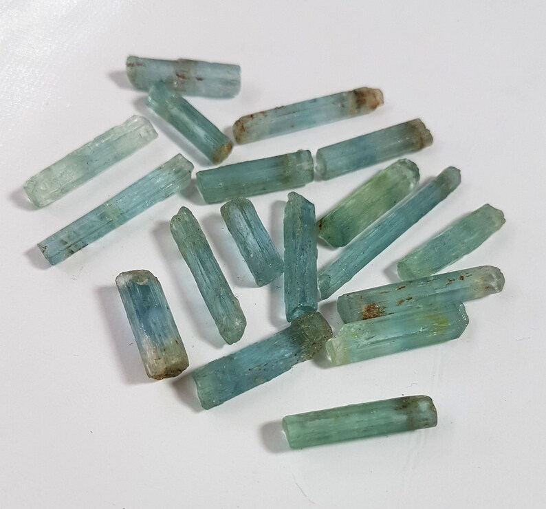 May include: A collection of raw aquamarine crystals, each with a distinct, elongated shape and a light blue-green color. The crystals are arranged on a white surface.