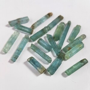 May include: A collection of aquamarine crystals, each with a distinct, elongated shape and a light blue to greenish-blue color. The crystals are arranged in a random pattern on a white surface.