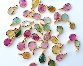 May include: A collection of gemstone pendants in various colors, including pink, green, and yellow, set in gold-colored metal. The pendants have a teardrop or oval shape and are scattered on a white surface. The gemstones appear to be tourmaline.