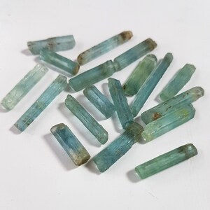 May include: A collection of light blue aquamarine crystals, each with a long, slender, and pointed shape. The crystals are arranged in a random pattern on a white surface.