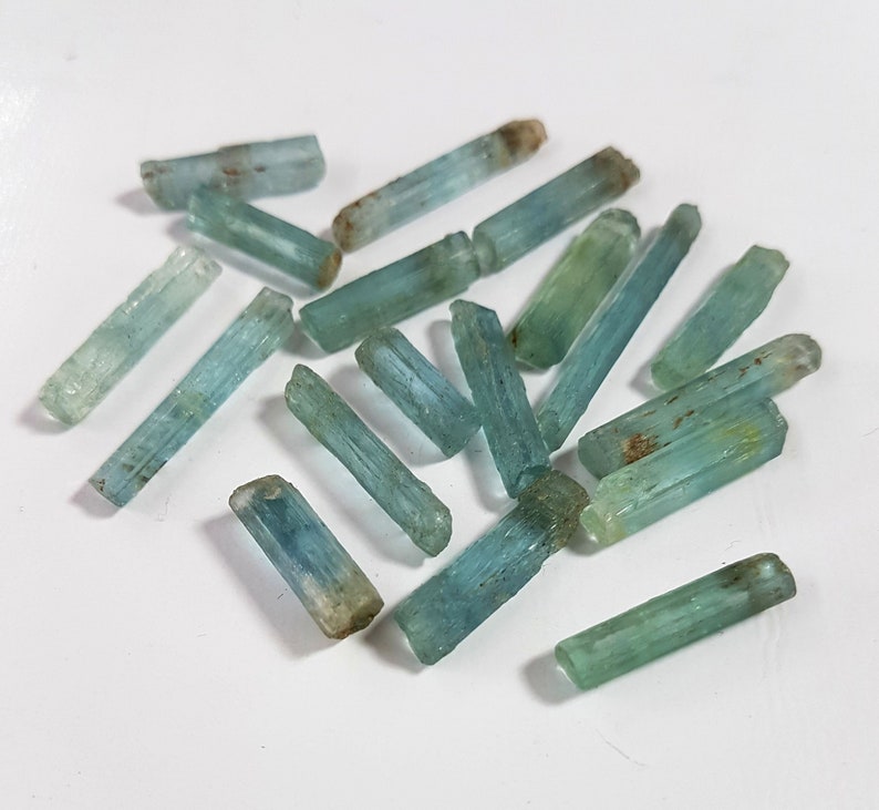 May include: A collection of light blue aquamarine crystals, each with a long, slender, and pointed shape. The crystals are arranged in a random pattern on a white surface.