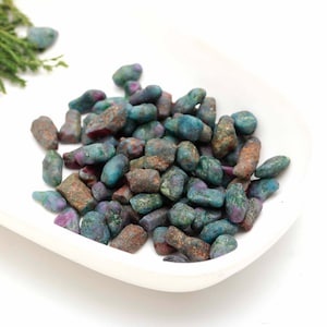 Puede incluir: Un montón de pequeñas piedras preciosas rugosas, verdes, azules y rojas. Las piedras preciosas están esparcidas en un plato blanco.