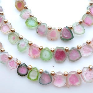 May include: A strand of watermelon tourmaline gemstone slices arranged in a row, featuring natural pink and green color gradients. Each translucent, irregularly shaped stone is separated by a small, polished gold-tone metal spacer bead.