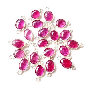 May include: A collection of 20 oval-shaped ruby red gemstone charms with silver jump rings. The charms are arranged in a circular pattern on a white background.