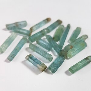 May include: A collection of light blue aquamarine crystals, each with a pointed tip and a smooth, polished surface. The crystals are arranged in a loose pile on a white background.
