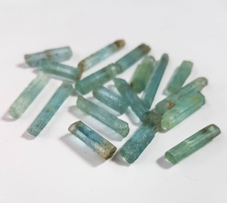 May include: A collection of light blue aquamarine crystals, each with a pointed tip and a smooth, polished surface. The crystals are arranged in a loose pile on a white background.