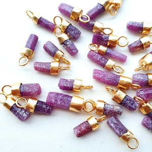 May include: A collection of raw ruby gemstone pendants. Each pendant features a cylindrical, rough-cut ruby stone with gold-colored metal caps and a small loop for attaching to a chain or other jewelry component. The rubies display a deep pink-purple hue.