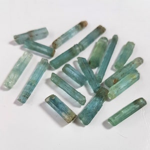 May include: A collection of aquamarine crystals, each with a distinct, elongated shape and a light blue to greenish-blue color. The crystals are arranged in a random pattern on a white surface.