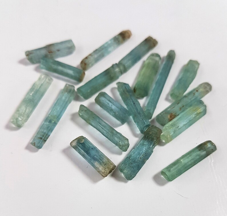 May include: A collection of aquamarine crystals, each with a distinct, elongated shape and a light blue to greenish-blue color. The crystals are arranged in a random pattern on a white surface.