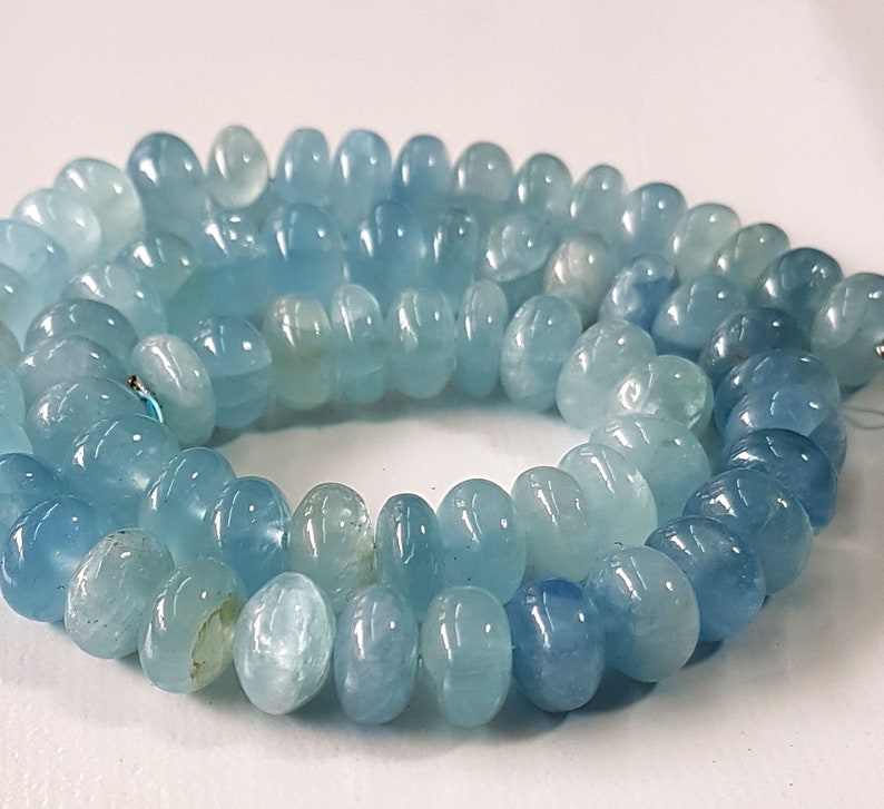 May include: A strand of light blue aquamarine gemstone beads. The beads are smooth and round, with a slightly flattened shape. They are strung on a clear thread.