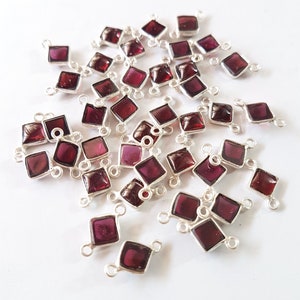 Pode incluir: Um grupo de conectores prateados com pedras preciosas vermelhas de forma quadrada. Os conectores têm dois laços de cada lado, permitindo que sejam anexados a correntes de joias ou outros componentes.