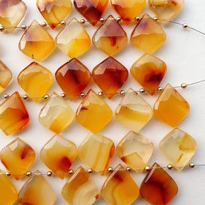 Puede incluir: Hilo de cuentas facetadas en forma de diamante en tonos ámbar, naranja y marrón. Las cuentas están ensartadas en un fino alambre plateado con pequeñas cuentas espaciadoras doradas. Las piedras preciosas tienen un aspecto pulido y translúcido.