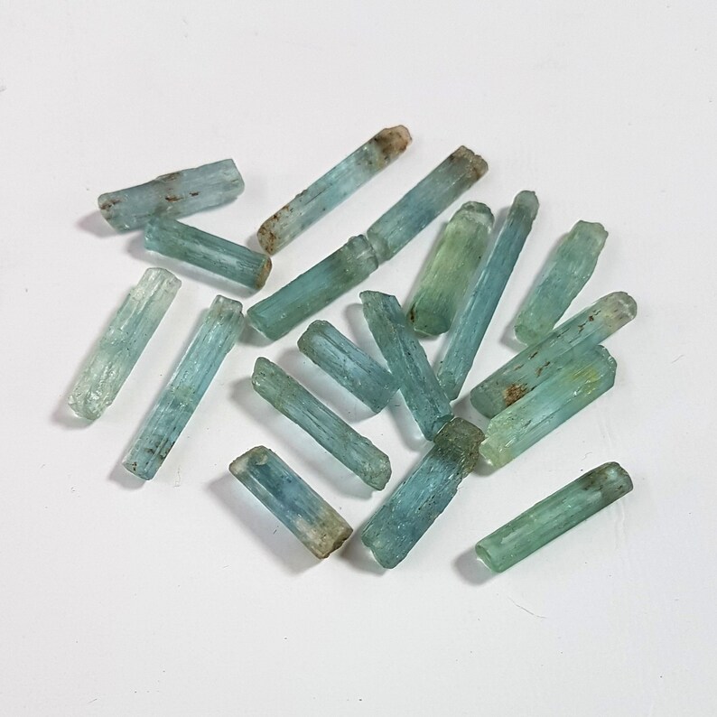 May include: A collection of light blue aquamarine crystals, each with a long, slender, and pointed shape. The crystals are arranged in a random pattern on a white surface.