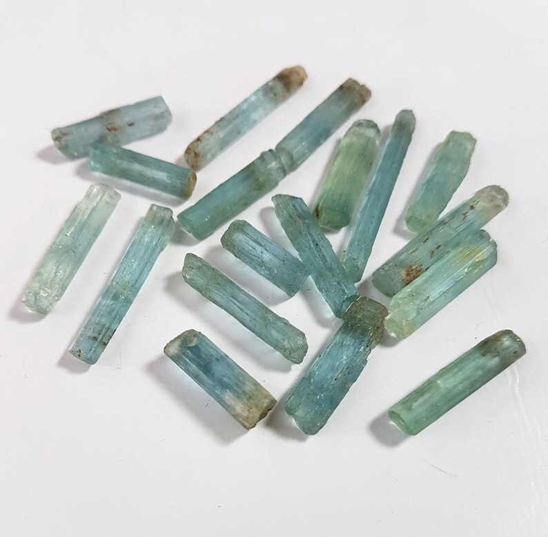 May include: A collection of light blue aquamarine crystals, each with a long, slender, and pointed shape. The crystals are scattered on a white surface.