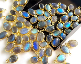 May include: A collection of oval-shaped labradorite gemstone pendants with gold-colored metal settings. The gemstones display iridescent blue and gray hues. The pendants are scattered on a white surface and in a black tray, ready for jewelry making.