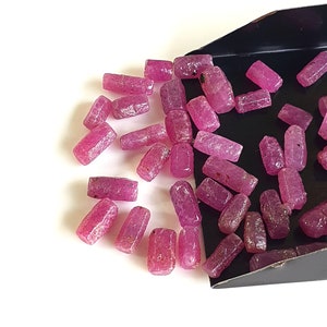 Op de afbeelding: Een verzameling roze robijn edelstenen, waarvan sommige in een zwarte lade liggen. De edelstenen zijn ruw en hebben een natuurlijke, onbewerkte uitstraling.