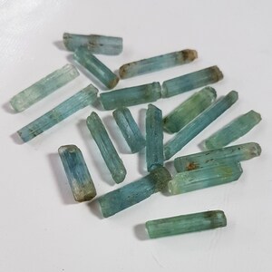 May include: A collection of light blue aquamarine crystals, each with a long, slender, and pointed shape. The crystals are arranged in a random pattern on a white surface.