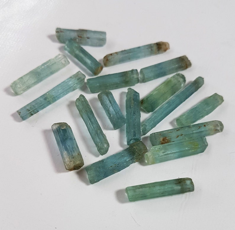 May include: A collection of light blue aquamarine crystals, each with a long, slender, and pointed shape. The crystals are arranged in a random pattern on a white surface.