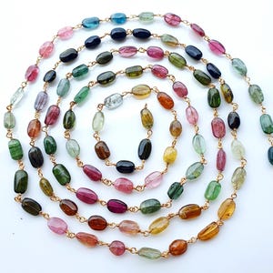 May include: A long chain featuring smooth, oval-shaped multi-colored tourmaline gemstones linked together with thin gold-toned wire. The stones display a variety of translucent hues including pink, green, blue, yellow, brown, and black.