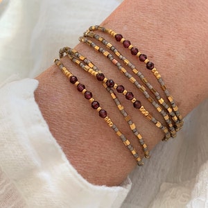 May include: A set of four delicate beaded bracelets featuring a combination of metallic gold, matte grey, and deep red garnet-coloured beads. The bracelets are arranged in a stacked formation, showcasing alternating patterns of small round and cylindrical beads.
