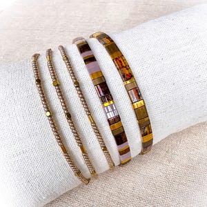 Può includere: Un set di cinque bracciali composto da tre fili sottili di piccole perline dorate metallizzate e due fili più larghi di tessere rettangolari piatte nei toni del marrone, bronzo e viola tenue, disposti su un tessuto bianco testurizzato.