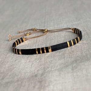 Peut inclure: Un bracelet en perles noires et dorées avec une fermeture en cordon marron. Le bracelet est composé de petites perles carrées avec des accents dorés.