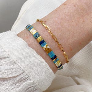Può includere: Due braccialetti su un polso. Uno è un braccialetto a catena d'oro. L'altro è un braccialetto di perline con perline blu, bianche, dorate e marroni. I braccialetti sono su un polso di carnagione chiara.