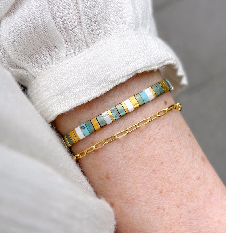 May include: A two-piece bracelet set featuring one strand of rectangular beads in alternating turquoise, white, and gold tones, paired with a second strand consisting of a delicate gold-toned metal chain with elongated oval links.