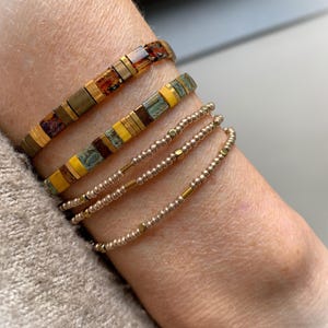 Può includere: Un set di quattro braccialetti composto da due fasce larghe realizzate con tessere rettangolari color ambra, marrone, giallo e verde acqua, e due fili sottili di piccole perline metalliche con distanziatori dorati in un design a strati.