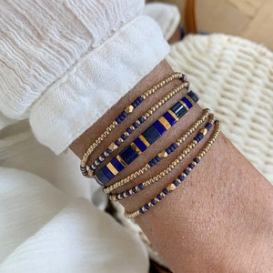 May include: A stack of five beaded bracelets featuring a combination of small metallic gold beads, matte navy blue seed beads, and a central row of rectangular blue lapis lazuli-style tiles interspersed with thin metallic gold spacers.