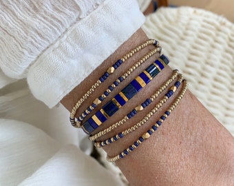May include: A stack of five beaded bracelets featuring a combination of small metallic gold beads, matte navy blue seed beads, and a central row of rectangular blue lapis lazuli-style tiles interspersed with thin metallic gold spacers.