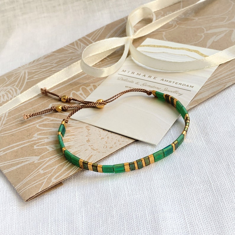 Puede incluir: Un brazalete de cuentas verdes y doradas con cierre de cord&oacute;n marr&oacute;n. El brazalete est&aacute; sobre una bolsa de papel marr&oacute;n con una etiqueta blanca que dice "Mirmare Amsterdam, Joyer&iacute;a delicada y de piedras preciosas, www."