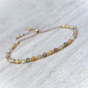 May include: A delicate adjustable bracelet featuring a series of small, faceted, multi-colored gemstone beads in shades of gray, tan, and peach, interspersed with tiny, polished gold-tone metal spacer beads on a thin, light-colored cord.